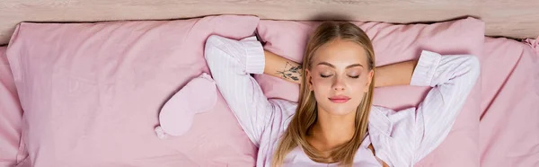 Вид зверху на молоду жінку в піжамі, що лежить біля маски на ліжку, банер — стокове фото