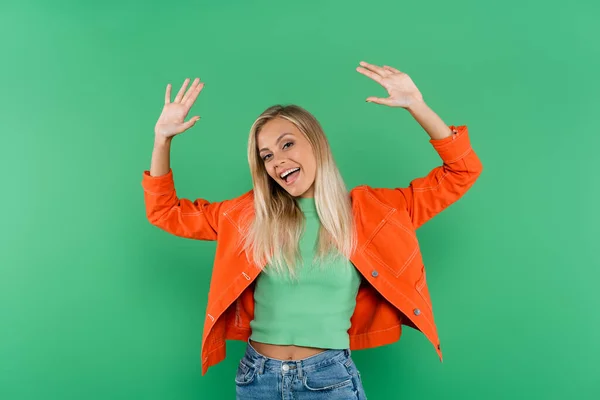 Mujer rubia excitada en jeans y chaqueta naranja agitando las manos aisladas en verde - foto de stock