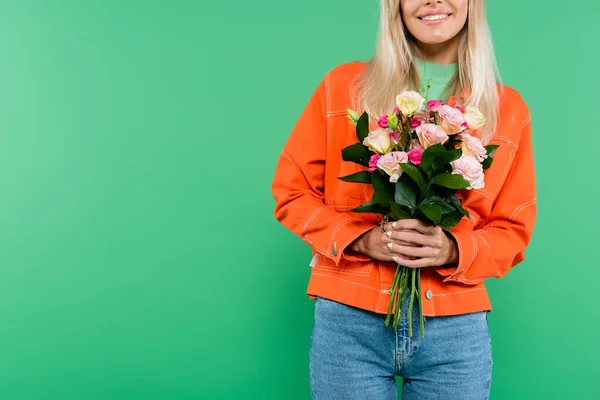 Cropped view of smiling woman in orange jacket and jeans holding flowers isolated on green — Stock Photo