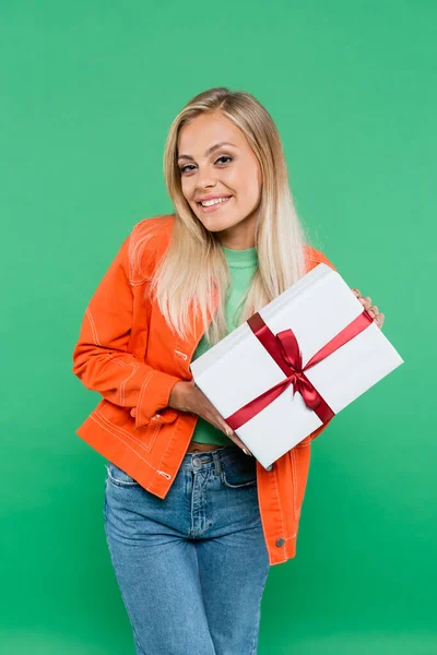 Fröhliche blonde Frau in orangefarbener Jacke hält Geschenk isoliert auf Grün — Stockfoto