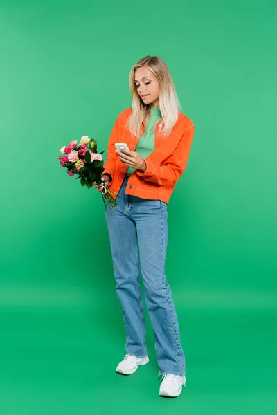 Ansicht der trendigen blonden Frau mit Blumenstrauß auf dem Smartphone in voller Länge auf grün — Stockfoto