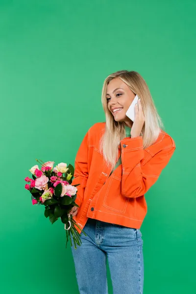Pleased woman in orange jacket holding bouquet while talking on mobile phone isolated on green — Stock Photo