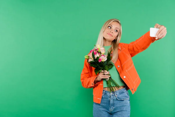 Pretty blonde woman with flowers taking sefie on mobile phone isolated on green — Stock Photo