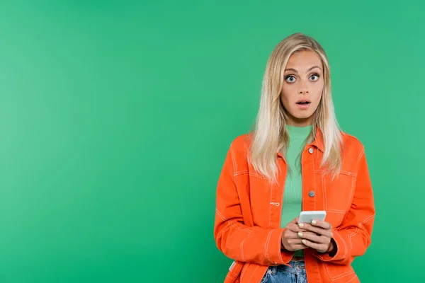 Femme blonde surprise en veste orange regardant la caméra tout en bavardant sur le téléphone portable isolé sur vert — Photo de stock