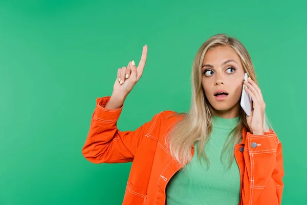 Amazed blonde woman pointing up with finger while talking on mobile phone isolated on green — Stock Photo