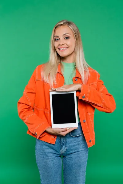 Mujer rubia alegre en jeans y chaqueta naranja mostrando tableta digital con pantalla en blanco aislado en verde - foto de stock