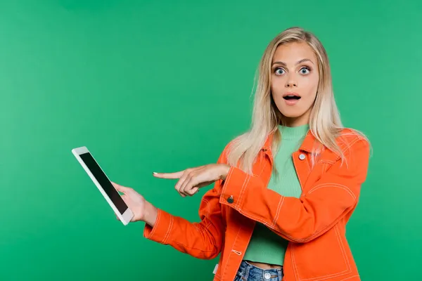 Erstaunte Frau in orangefarbener Jacke zeigt auf digitales Tablet mit leerem Bildschirm auf grünem Hintergrund — Stockfoto