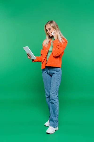 Femme souriante et à la mode parlant sur un téléphone mobile tout en utilisant une tablette numérique sur vert — Photo de stock