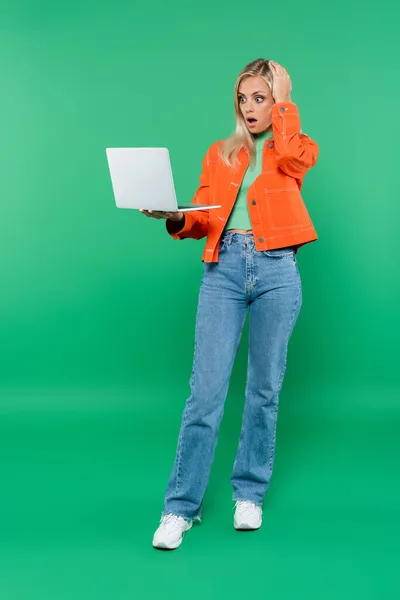 Mulher chocada em roupas elegantes tocando cabeça enquanto olha para laptop no verde — Fotografia de Stock