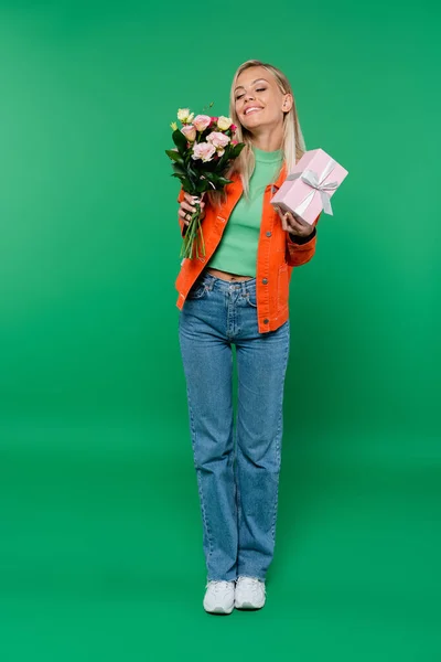 Full length view of happy blonde woman with closed eyes holding bouquet and gift box on green — Stock Photo