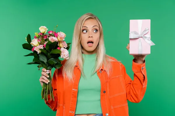Erstaunt blonde Frau in orangefarbener Jacke mit Geschenkbox und bunten Blumen isoliert auf grün — Stockfoto