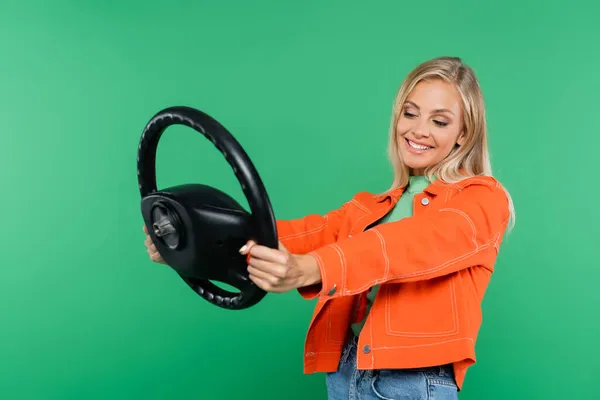 Mujer joven y feliz en chaqueta naranja posando con volante aislado en verde - foto de stock