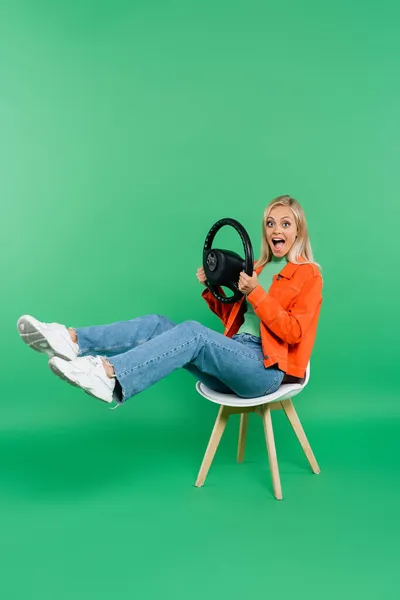 Mujer asombrada con volante mirando a la cámara mientras está sentado en la silla con las piernas levantadas en verde - foto de stock