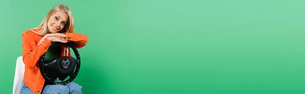 Jovem loira em jaqueta elegante sentado na cadeira com volante e olhando para a câmera isolada no verde, banner — Fotografia de Stock