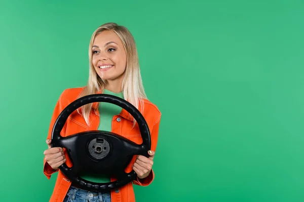 Mujer rubia positiva con chaqueta naranja mirando hacia otro lado mientras sostiene el volante aislado en verde - foto de stock