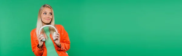 Mulher loira bonita em roupas da moda segurando fones de ouvido enquanto olha para longe isolado no verde, banner — Fotografia de Stock