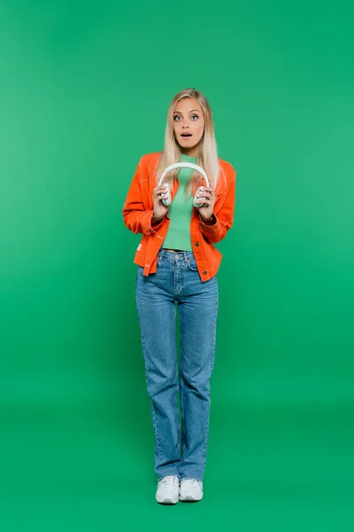 Full length view of amazed woman in orange jacket and jeans holding headphones on green — Stock Photo