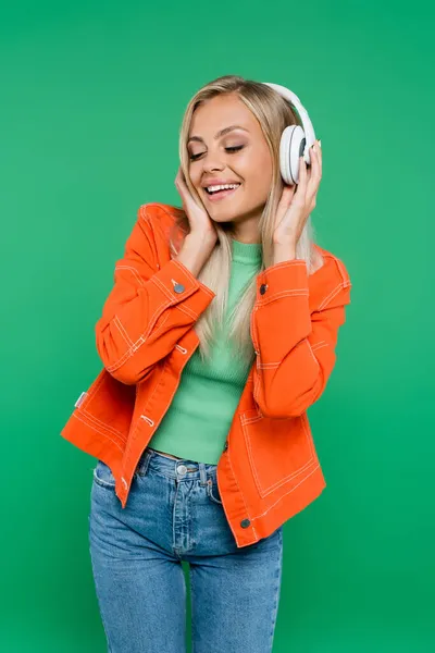 Cheerful woman in orange jacket and headphones listening music with closed eyes isolated on green — Stock Photo
