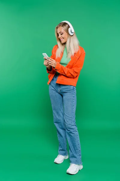 Full length view of happy woman in headphones using smartphone on green — Stock Photo