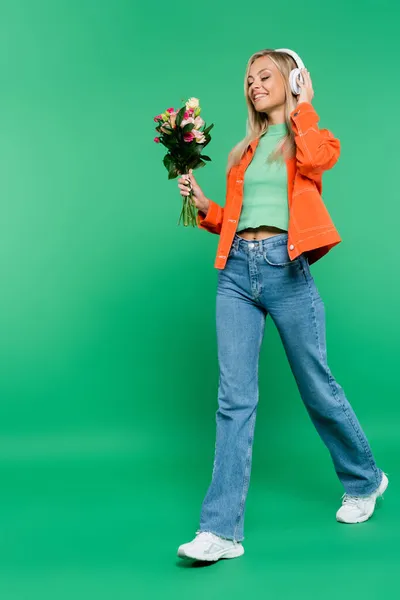 Full length view of cheerful blonde woman in headphones, orange jacket and jeans walking with bouquet on green — Stock Photo