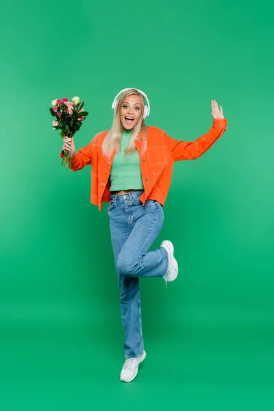 Excited woman in headphones and trendy clothes posing with flowers on green — Stock Photo