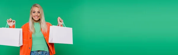 Happy blonde woman in trendy clothes looking at camera while showing shopping bags isolated on green, banner — Stock Photo