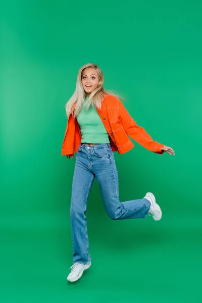 Visão de comprimento total da mulher loira em jeans e jaqueta de laranja sorrindo enquanto corre em verde — Fotografia de Stock