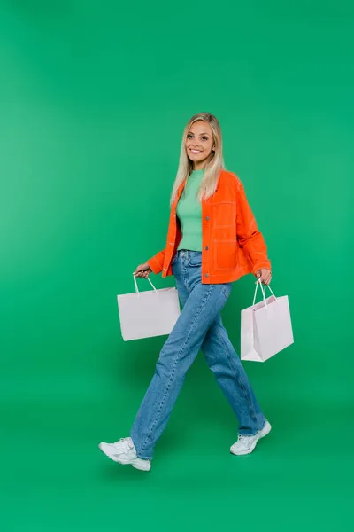 Улыбающаяся женщина в оранжевой куртке и джинсах ходит с пакетами покупок на зеленом — стоковое фото