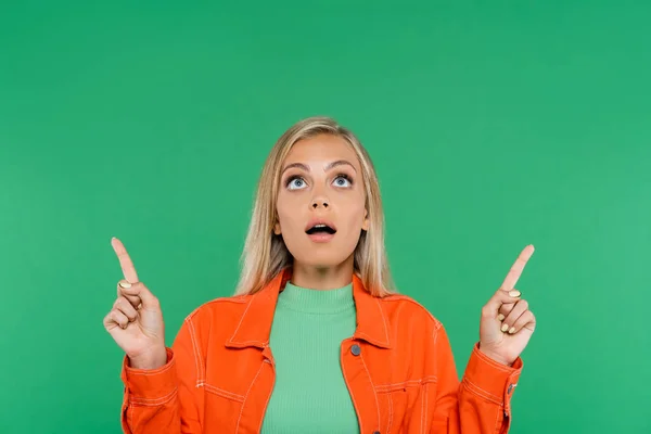 Mujer rubia asombrada mirando hacia arriba y señalando con los dedos aislados en verde - foto de stock