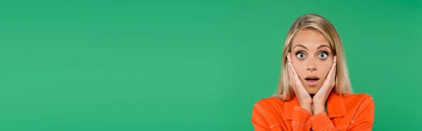 Astonished blonde woman touching face while looking at camera isolated on green, banner — Stock Photo