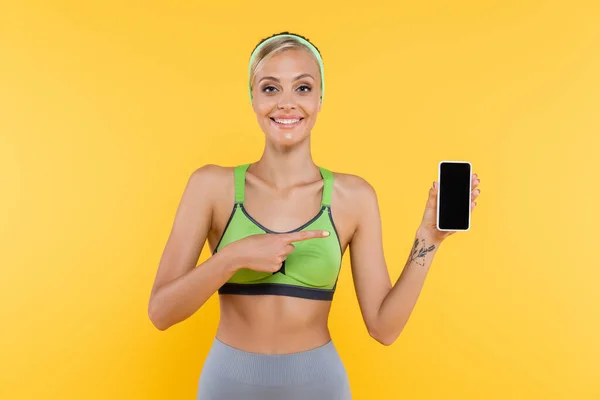 Mulher alegre em sportswear apontando para o telefone celular com tela em branco isolado no amarelo — Fotografia de Stock