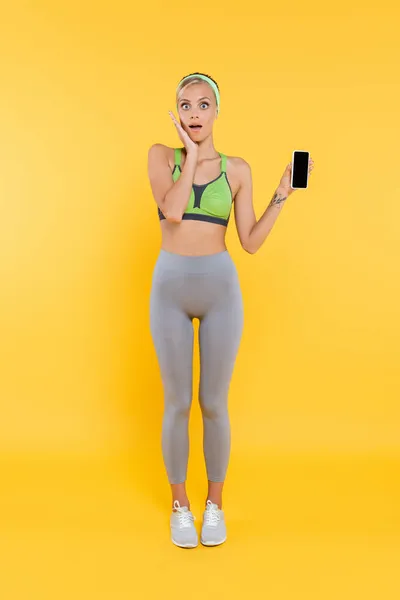 Vista completa de la mujer sorprendida en la cara de tocar ropa deportiva mientras que muestra el teléfono inteligente con pantalla en blanco en amarillo - foto de stock