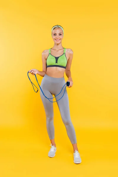 Vue pleine longueur de la femme mince en vêtements de sport debout avec corde à sauter sur jaune — Photo de stock