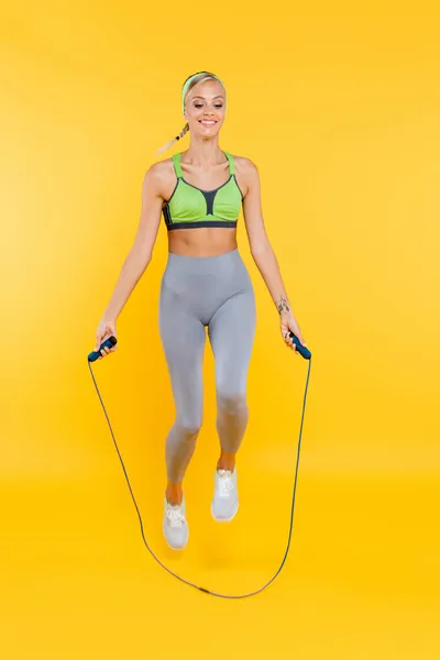 Vista a figura intera di donna felice in leggings grigi e reggiseno sportivo verde che salta con corda da salto sul giallo — Foto stock