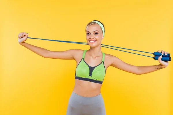 Joyeuse jeune femme souriant à la caméra tout en s'entraînant avec corde à sauter isolé sur jaune — Photo de stock