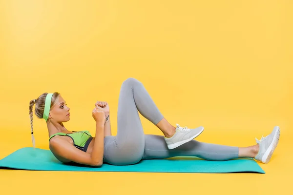 Vista laterale della donna in leggings grigi e reggiseno sportivo verde che esercita sul tappeto fitness sul giallo — Foto stock