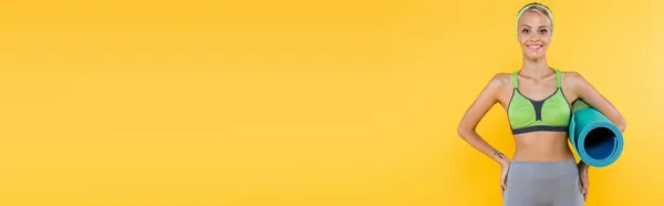 Donna magra sorridente alla macchina fotografica mentre in piedi con le mani sui fianchi e tappetino fitness isolato su giallo, banner — Foto stock