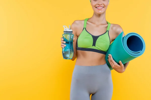 Vista recortada de la mujer sonriente en ropa deportiva celebración de botella deportiva y alfombra de fitness aislado en amarillo - foto de stock