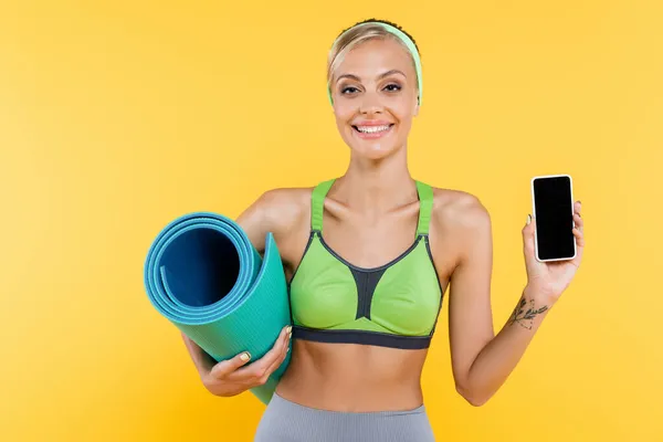 Mulher feliz em sutiã de esportes verde segurando tapete de fitness ao mostrar o telefone móvel com tela em branco isolado no amarelo — Fotografia de Stock