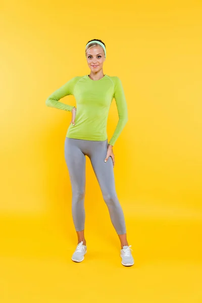Vista a figura intera di bella donna in leggings sportivi e t-shirt a maniche lunghe in piedi con mano sul fianco su giallo — Foto stock
