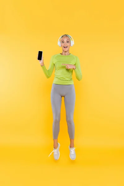 Full length view of amazed sportswoman in headphones pointing at cellphone with bank screen on yellow — Stock Photo