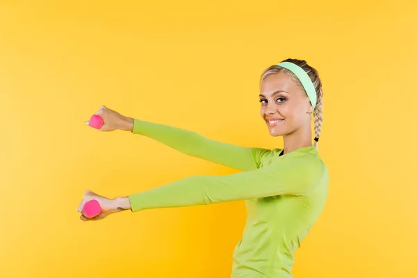 Задоволена жінка посміхається на камеру під час тренувань з гантелями ізольовані на жовтому — стокове фото