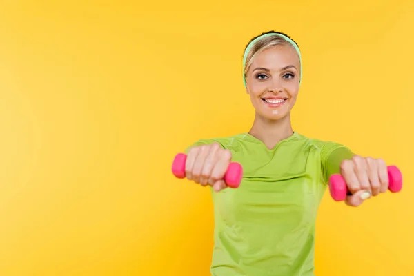 Slim woman in green long sleeve shirt smiling while working out with dumbbells isolated on yellow — Stock Photo