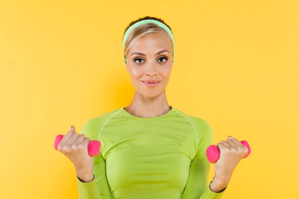 Sportive woman exercising with dumbbells while smiling at camera isolated on yellow — Stock Photo