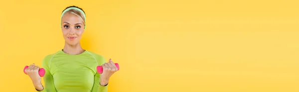 Pretty and smiling woman working out with dumbbells while looking at camera isolated on yellow, banner — Stock Photo