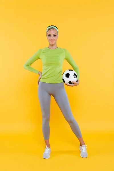 Vista integrale della sportiva in piedi con mano sul fianco e pallone da calcio sul giallo — Foto stock