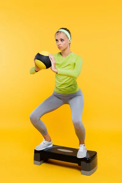 Woman in sportswear holding ball while doing sit ups on step board on yellow — Stock Photo