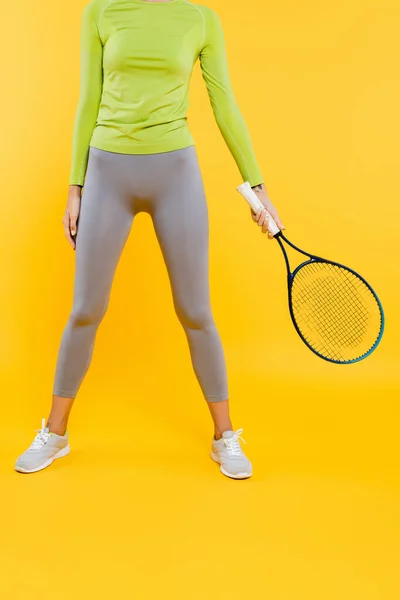 Cropped view of woman in sports leggings and sneakers holding tennis racquet on yellow — Stock Photo
