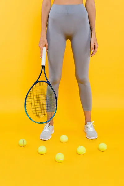 Partial view of sportswoman in grey leggings holding racquet while standing near tennis balls on yellow — Stock Photo