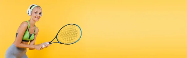 Усміхнена спортсменка в навушниках грає в теніс ізольовано на жовтому, банер — стокове фото